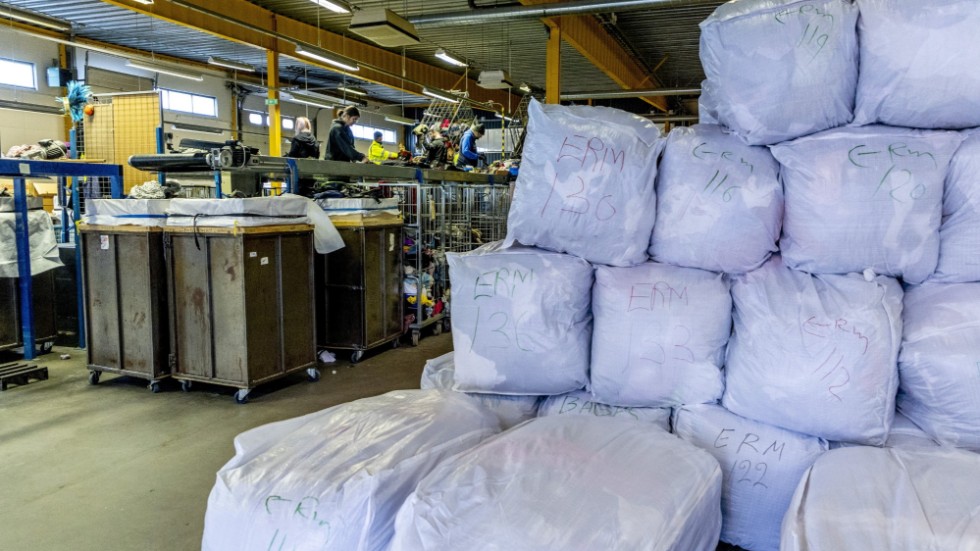 Stora klädsäckar som väntar på export från Myrornas sorteringsanläggning i Göteborg. I bakgrunden sorterar anställda kläder vid två löpande band.