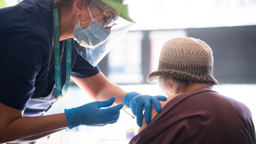 En äldre dam i hatt blir vaccinerad mot covid-19 av en sköterska i visir och ansiktsskydd.
