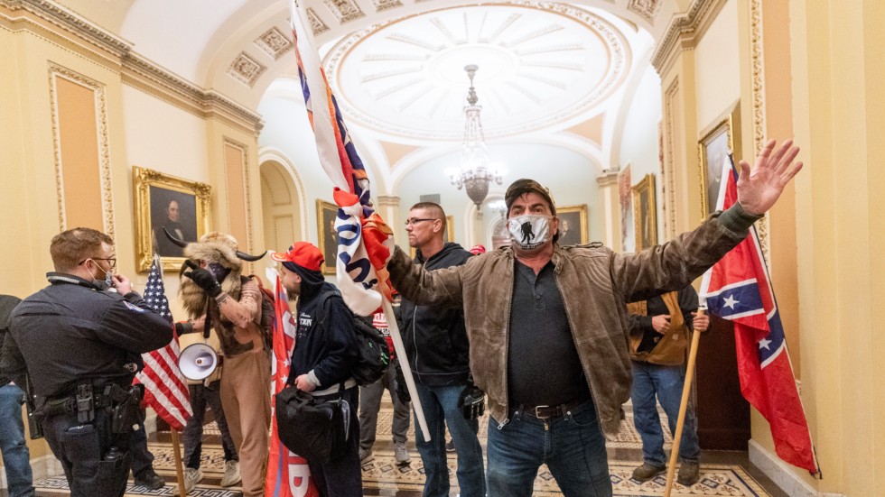 Trumpanhängare som brutit sig in i kongressbyggnaden möter en ensam polisman i en hall nära senatens plenisal. Till höger i bild en sydstatsflagga, använd som symbol av rasistiska högerextremister.