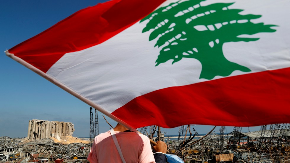 En man med den libanesiska flaggan tittar ut över förödelsen i hamnen efter tisdagens explosion i Beirut.