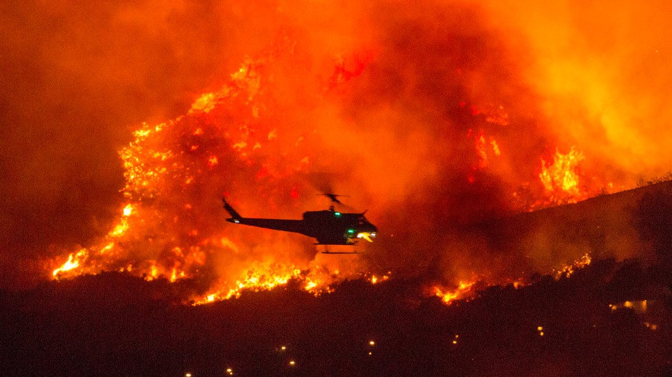 En brandhelikopter på väg att släppa vatten över en skogsbrand i Yucaipa i Kalifornien. I en annan helikopterinsats har tiotals människor räddats undan lågorna.